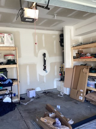 A disorganized garage interior showing bare drywall, a concrete floor with scattered boxes and items, and open shelving on the sides, before the installation of overhead storage racks.