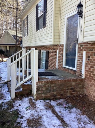 Before photo of a front porch renovation project. The existing porch has a worn-out wooden railing and steps, with a concrete base and brick accents.