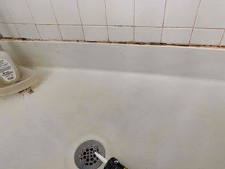 A bathtub area with visible mold and mildew buildup along the grout lines between the tiles and the tub. The caulk around the edge of the tub appears damaged and discolored, with gaps and areas where the sealant has worn away. 
