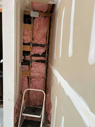 A narrow space with exposed wooden framing and insulation. The drywall is being prepared, with visible joints that have been taped and are being smoothed with joint compound. 