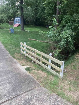  The image shows a newly repaired wooden fence along the edge of a driveway. The fence consists of white-painted posts with decorative caps and fresh, unpainted horizontal wooden slats. The surrounding area features a grassy yard with a small blue shed, trees, and a well structure in the background.