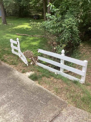 The image shows the fence before repairs, with significant damage. A section of the fence is broken and leaning, with cracked and splintered wooden slats. The paint on the remaining portion of the fence is chipped and worn. 