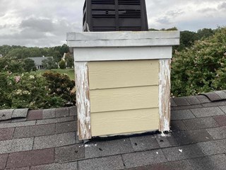 The image shows a damaged exterior chimney trim before repairs. The chimney structure has beige siding with white trim that is peeling and deteriorating, exposing bare wood. The top trim appears weathered, and there are visible signs of water damage and wear.