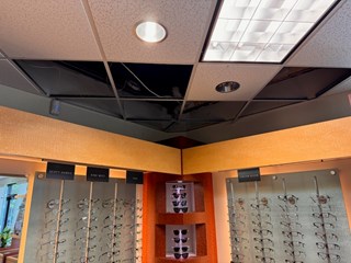  The image shows a damaged drop ceiling in a commercial eyewear store. Several ceiling tiles are missing, exposing the dark space above with visible pipes and wiring. The surrounding ceiling has recessed lighting, a large fluorescent light panel, and a partially visible air vent.