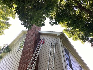 The image shows the same house with a repair in progress. A tall extension ladder is propped against the house, reaching up to the fascia area near the chimney. The trim appears to have been repainted or replaced, looking fresher and cleaner.