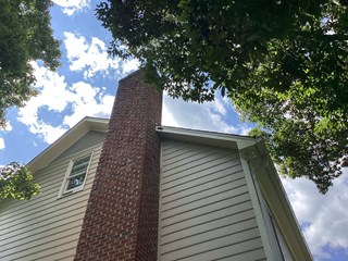  The image shows the exterior of a house with a tall brick chimney and beige siding. The fascia and trim near the roofline appear weathered and in need of maintenance. 