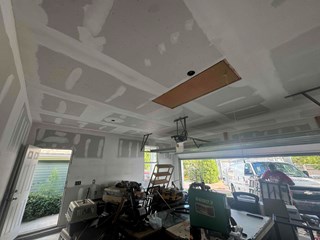 The image shows the garage after insulation and drywall installation. The walls and ceiling are now covered with white drywall, with visible joint compound applied to the seams. The overall space appears more finished and enclosed compared to the first image.
