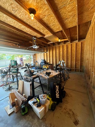 The image shows an unfinished garage with exposed wooden framing and ceiling beams. The space is cluttered with woodworking tools, power equipment, and construction materials. The walls lack insulation and drywall, exposing the wooden structure.