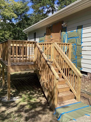 The image shows an unfinished wooden deck attached to a house, with stairs leading down to the yard. The deck appears to be newly built, with raw, unstained wood. The house's exterior wall is covered with protective paper and tape, suggesting preparation for painting or staining.