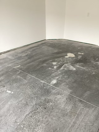 The image shows a room with unfinished concrete flooring and white walls. The baseboards are not yet installed, leaving a gap between the wall and the floor.