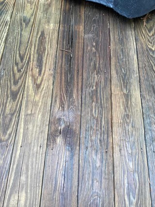  The image shows a close-up of an aged wooden deck with visible discoloration, cracks, and signs of wood rot. The planks appear weathered, with some areas darker than others, and there is a noticeable split in one of the boards. The wood grain is prominent, and some nails are slightly raised.