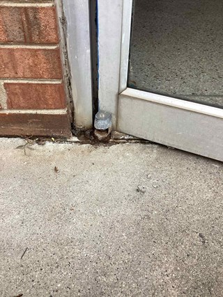Close-up of the bottom corner of a commercial glass door with an aluminum frame. The door hinge is rusted and corroded, with visible dirt and debris accumulated around the base. 