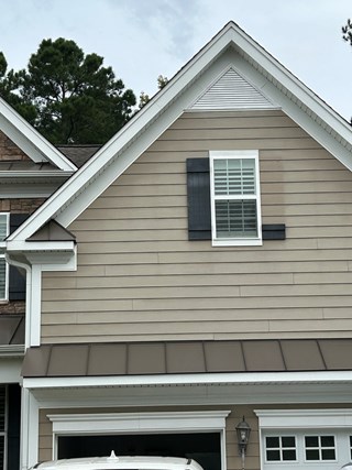 Exterior of a house with beige siding, white trim, and a single window with dark gray shutters. One of the shutters is partially damaged, with a large portion missing. 