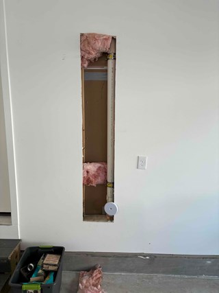 Section of drywall cut out from a white interior wall, revealing insulation and a PVC pipe inside. Pieces of pink fiberglass insulation are partially removed.