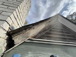 Damaged fascia and soffit area near the roofline of a house. The wooden fascia board is heavily weathered, with visible rot and peeling paint. A section of the trim is deteriorated, and the underlying structure is partially exposed.