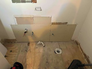 A bathroom under renovation with exposed plywood flooring and partially installed drywall. 