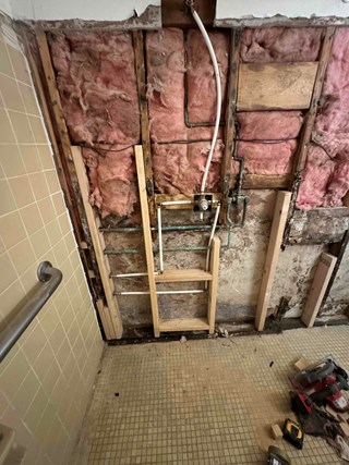 A damaged commercial shower wall with missing tiles exposing insulation, wooden framing, and plumbing. 