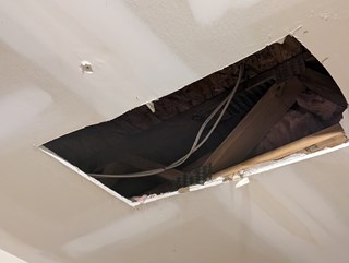 A section of a ceiling with a large rectangular hole, exposing wooden beams, insulation, and electrical wiring