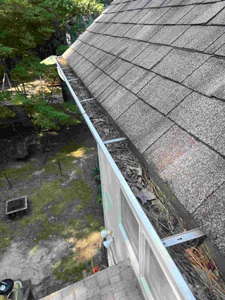 Shingle gutter system clogged with leaves, twigs, and pine needles. The buildup is heavy, and the debris obstructs the water flow.
