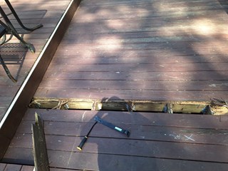A weathered wooden deck with several damaged boards removed, exposing the underlying wooden frame and joists. Some boards show signs of rot and decay.