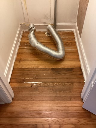  A small utility closet with damaged hardwood flooring. The wood is scuffed, stained, and uneven in some areas