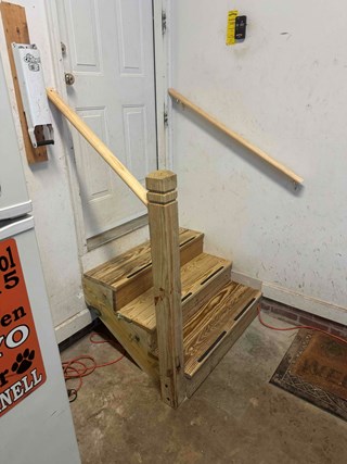 Garage entrance after repair, featuring newly constructed wooden steps with smooth, finished boards. A sturdy handrail has been installed on the left side, along with a wall-mounted handrail on the right for added safety. 