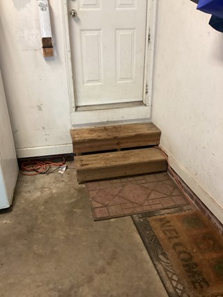 A garage entrance with two makeshift wooden steps leading to a white door. The steps are uneven, unfinished, and lack railings.
