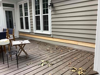 The side of a house with partially replaced siding. A section of the lower wall is stripped, and a new wooden trim board is being added along the base of the house. 