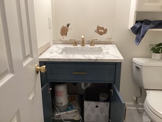 A bathroom with a vanity that is partially installed. The countertop is already in place with a marble finish, and there is a gold faucet. The wall behind the vanity shows some unfinished areas where the paint has been stripped or damaged, and there is an exposed patch of drywall.