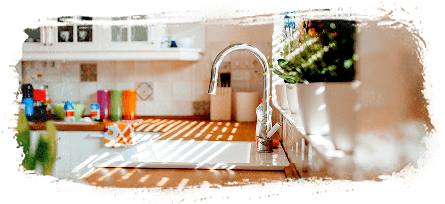 A farmhouse style kitchen sink with chrome hardware and a tan countertop. Sunlight shines through the window above the sink. A farmhouse style kitchen sink with chrome hardware and a tan countertop. Sunlight shines through the window above the sink.