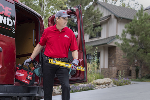 A uniformed Ace Handyman Craftsman holds a level in one hand and lifts a tool bag out of the back of a work van with the other.
