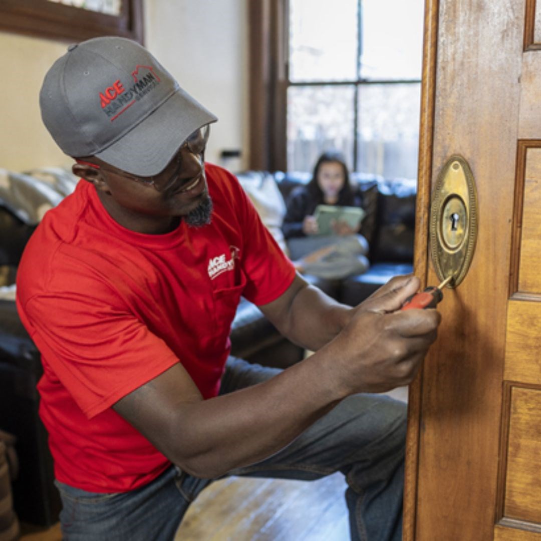 A man in an Ace Handyman Services ballcap and T-shirt fixes an interior sliding door while a woman on a couch in the background looks at a tablet. 