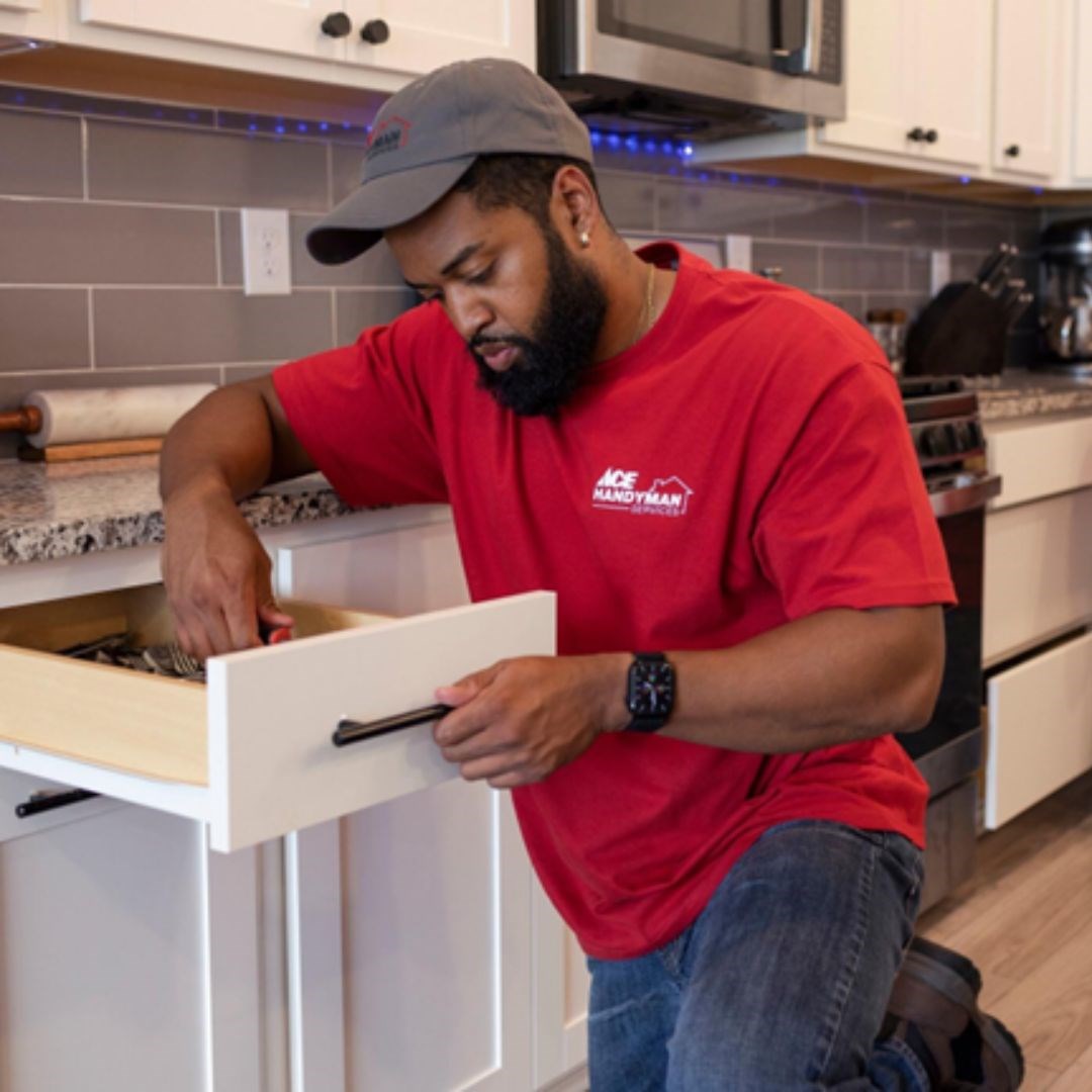 A man in Ace Handyman Services T-shirt and baseball cap kneels on the floor of a kitchen and perform repairs on drawers below the kitchen countertop. A man in Ace Handyman Services T-shirt and baseball cap kneels on the floor of a kitchen and perform repairs on drawers below the kitchen countertop.