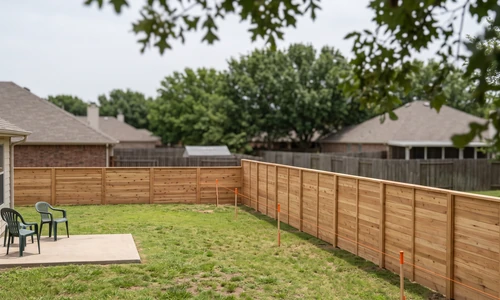 Elevated view of a new cedar fence line running along the rear of a suburban backyard with orange-capped survey stakes and a taut string line set inside it