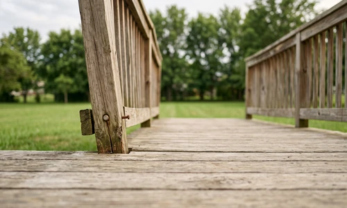 Wooden deck railing with a visibly loose post pulling away from the deck rim, gap open at the base, natural daylight