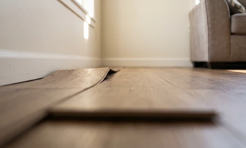 Section of laminate flooring lifting and gapping near a wall in a sunlit living room