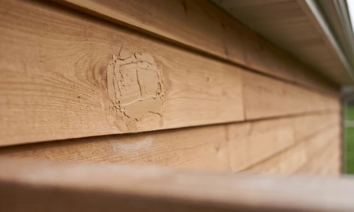 Rot pocket in a cedar siding board packed with tan wood-toned epoxy filler, feathered into the sound grain around it