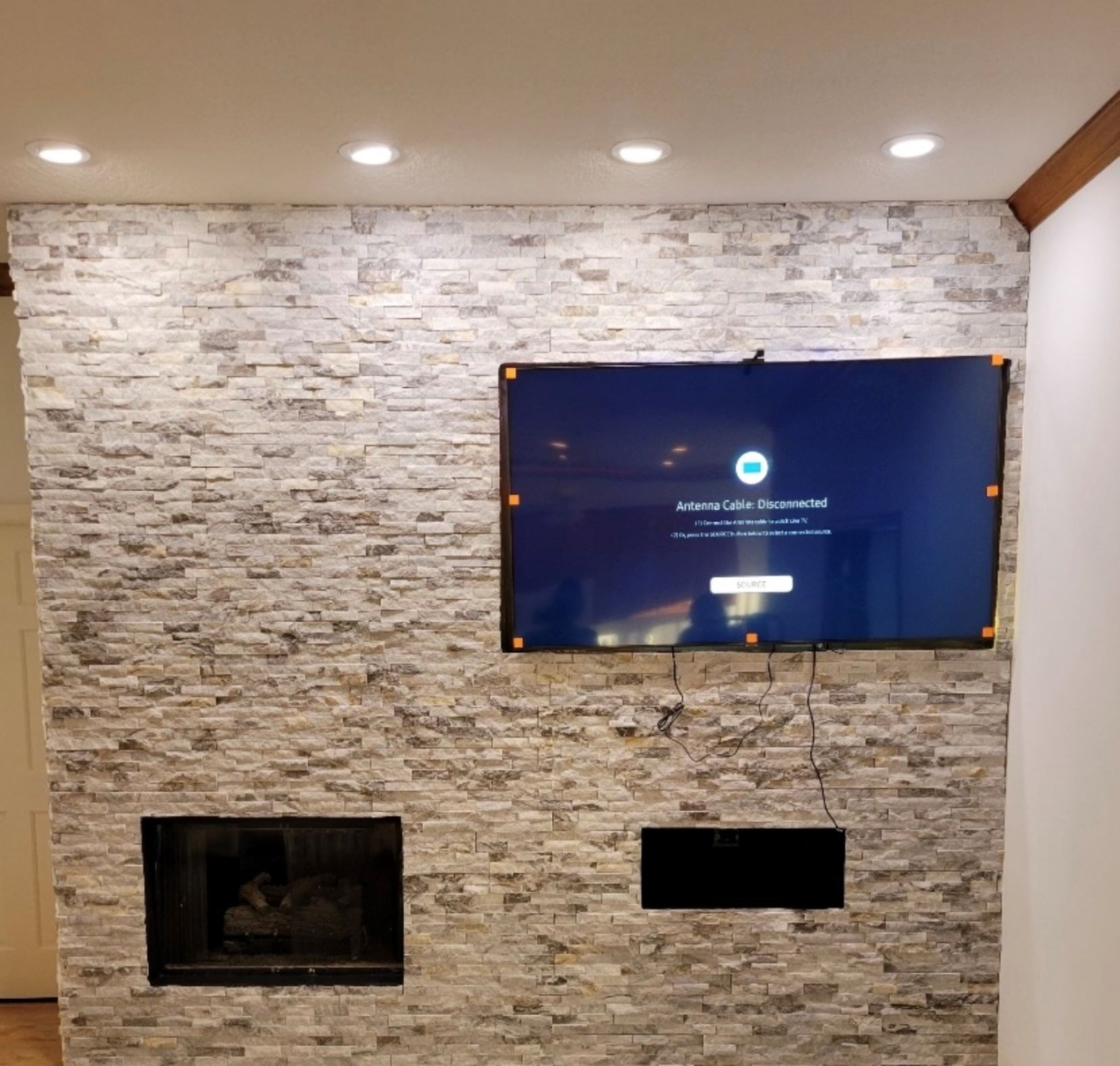 Mounted TV on stone wall with exposed wires, under ceiling lights.