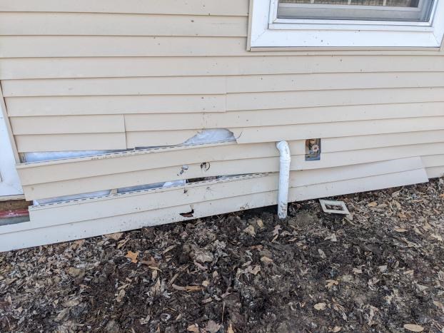 Damaged house siding near ground with exposed pipes.