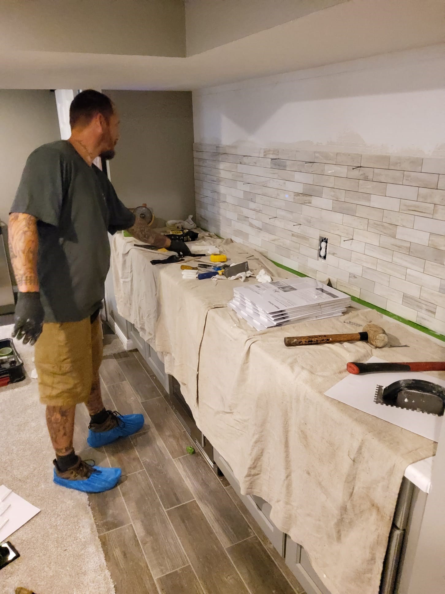 Handyman tiling a backsplash, tools on covered counter.